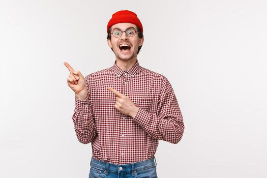 Charismatic funny young man in glasses and red beanie introduce something awesome, laughing and smiling excited pointing fingers upper left corner, recommend click or subscribe.