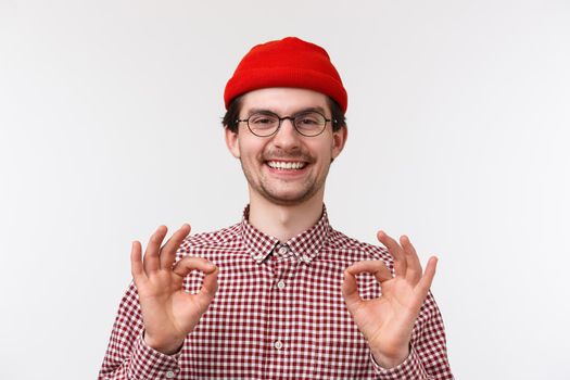 Close-up portrait caucasian male in glasses and red beanie, showing okay gesture rate good movie, being fully satisfied, guarantee all be perfect, standing pleased standing white background.