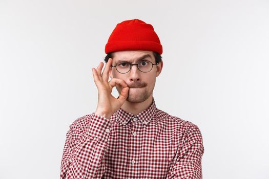 Close-up portrait of serious young caucasian guy with moustache, wear glasses and beanie, seal lips with finger gesture as if hiding secret, promise not speak about it, stand white background.