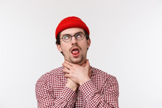 Close-up portrait of funny bearded guy chocking himself stick tongue as if cant breath, roll eyes pretend dying from boredom or express his distress, standing in red beanie and glasses.