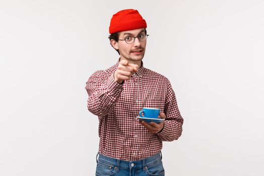 Waist-up portrait of handsome cheerful caucasian bearded man in glasses and red beanie, holding cup of coffee or tea and pointing at camera as telling to try this awesome drink yourself.