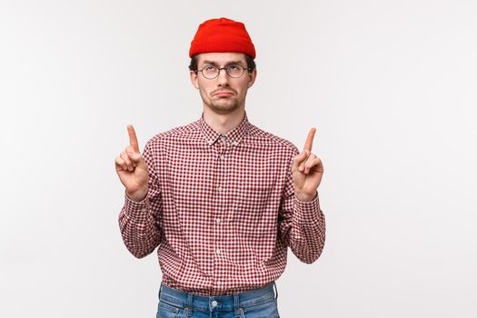 Waist-up portrait upset and disappointed whining adult man in red beanie, glasses, pointing fingers up at advertisement and showing dislike and uneasy feelings with face expression, white background.