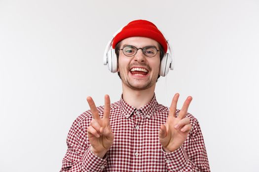 Music, technology and people concept. Positive and happy smiling young man with beard and glasses, wear beanie listen music in headphones, showing peace gesture and laughing, white background.