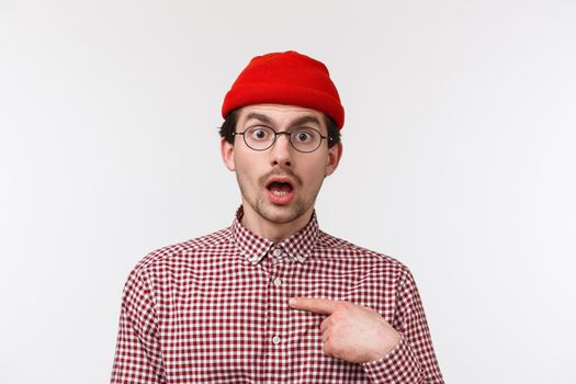 Close-up portrait surprised young hipster guy in glasses and beanie staring with disbelief, pointing himself indecisive, dont know if person for real mentioned him, standing white background.