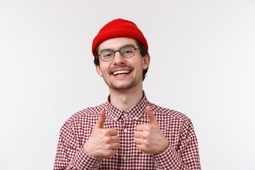 Nice work, good job mate. Supportive cute caucasian male in glasses and red beanie, show thumbs-up pleased, like or approve something awesome, smiling satisfied, standing white background.
