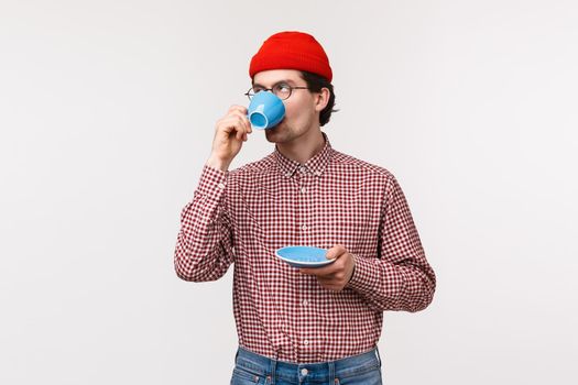 Skeptical and snobbish funny young man in red beanie and glasses, drinking coffee, sip tea from cup and roll eyes annoyed, listening to boring story, or feel satisfaction trying best drink ever.