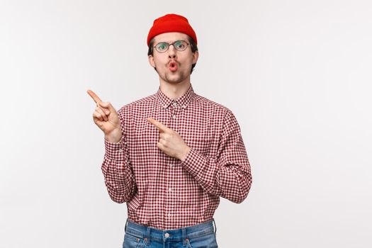 Waist-up portrait impressed and intrigued young caucasian bearded man in glasses, red beanie, pointing upper left corner and look camera curious, asking question about product, white background.