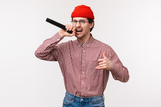 Funny and passionate cool hipster guy in red beanie and glasses singing rap song acting excited, holding microphone, having fun at karaoke with friends, standing white background joyful.