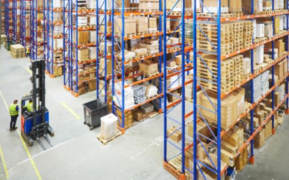 Blur warehouse background. Above view of warehouse workers moving goods and counting stock in aisle between rows of tall shelves full of packed boxes.