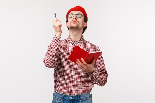 Waist-up portrait of funny male scientist working on project, passionatly look up and raise pen as have perfect idea, holding notebook, writing down his inspiring thoughts, stand white background.