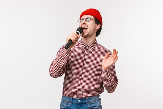 Brokenhearted guy singing romantic song in karaoke with passionate feeling, close eyes and scream into microphone, standing in red beanie, glasses and checked shirt, white background.