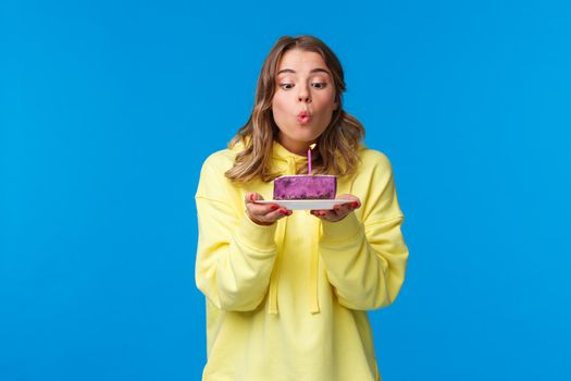Celebration, party and lifestyle concept. Silly cute european blond woman in yellow hoodie, making wish holding fruit cake blowing out b-day candle, standing blue background at birthday party.