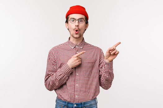 Surprised and curious funny bearded man in red beanie, glasses folding lips from amazement and interest asking question about cool new product in stock, pointing upper right corner.