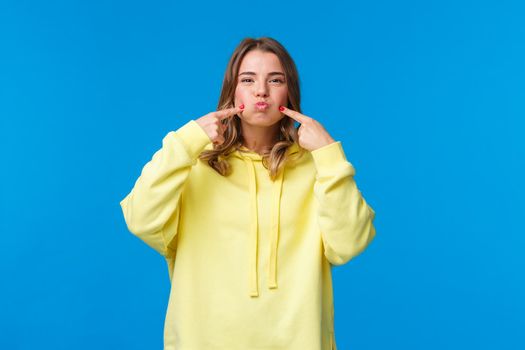 Women, lifestyle and emotions concept. Portrait of silly carefree girl fool around, hold her breath and pouting, poke cheeks trying not laugh, showing cute smile, look camera, blue background.