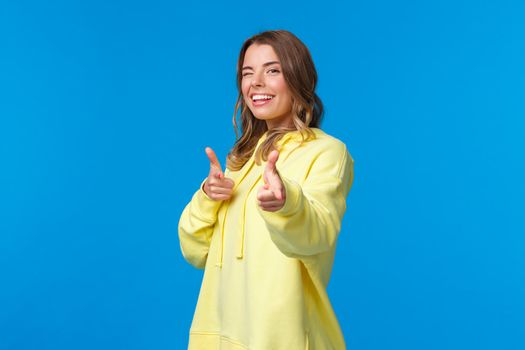 Hey you. Cheeky and carefree cute modern european girl in yellow hoodie, wink and make finger pistols gesture at camera to say hi, informal greeting concept. blue background.