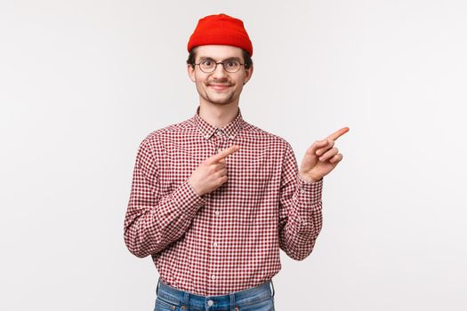 Waist-up portrait of satisfied happy smiling young hipster guy in red beanie, glasses pointing fingers right top advertisement with pleased expression, found excellent product or company service.