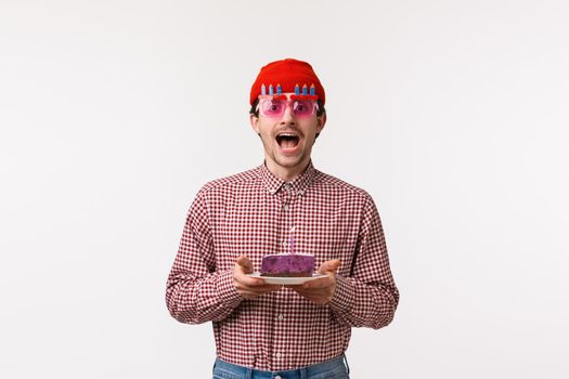 Celebration, holidays and lifestyle concept. Excited happy handsome bearded adult guy celebrating birthday with friends in funny glasses, holding b-day cake with lit candle, make wish.