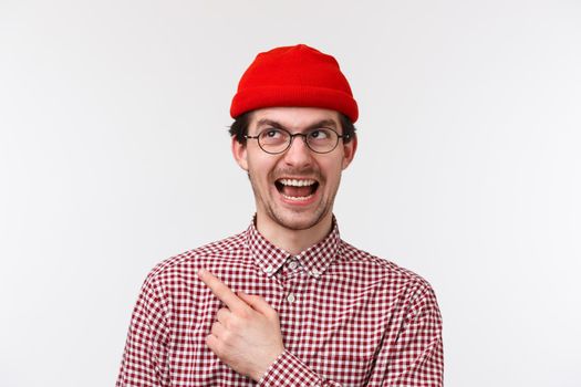 Close-up portrait cheerful handsome caucasian hipster guy in red beanie and glasses, pointing finger upper left corner and smiling pleased, showing advertisement, recommend product, special offer.