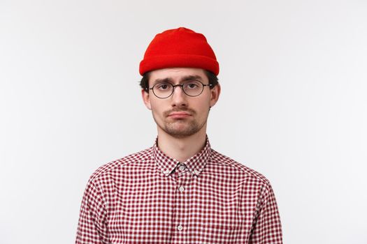 Close-up portrait of bored and reluctant young hipster guy feeling like loser, having bad unlucky day, standing in glasses and red beanie, feel lonely or distressed, stand white background.