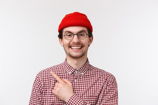 Close-up portrait cheerful handsome caucasian hipster guy in red beanie and glasses, pointing finger upper left corner and smiling pleased, showing advertisement, recommend product, special offer.