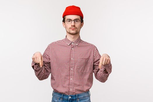 Waist-up portrait serious-looking determined young man in checked shirt, red beanie and glasses looking strict camera as pointing fingers down as something important and worth attention.