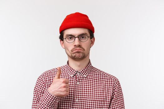Not bad, really good job. Close-up portrait satisfied serious-looking bearded young man in beanie and glasses make judgement, show thumb-up in approval or like gesture, white background.