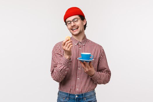 Waist-up portrait of funny caucasian hipster guy in red beanie, glasses and checked shirt, enjoying tasty cookie, eating dessert and drinking coffee or tea from blue cup on plate, white background.