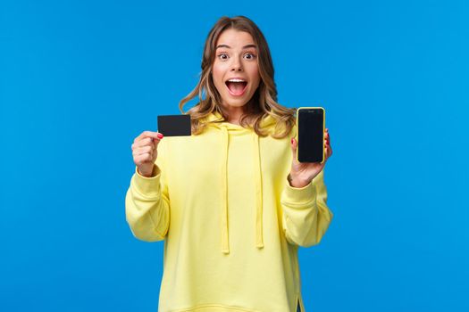 Amused and excited blond girl recommend using banking service, showing smartphone display and credit card and looking camera fascinated as receive bonus to bank account, blue background.