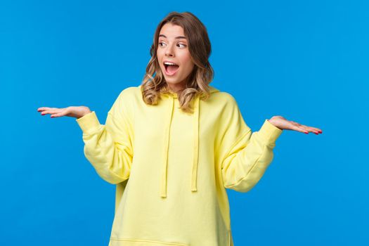 Girl weighing two choices as making decision, look away picking between products she holding in arms on left and right side, standing excited over blue background.