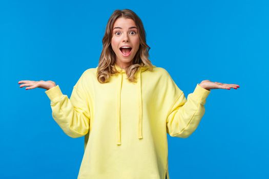 Upbeat good-looking blond caucasian female in yellow hoodie making choice, holding two products in hands on left and right side copy space, weighing them and look camera amused.