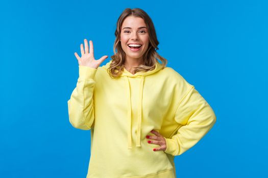 Cheerful friendly outgoing blond girl waving hand, informal greeting concept, smiling white teeth saying hi, welcoming new members to company, see friend, stand blue background.