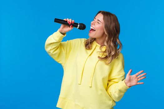 Carefree happy blond european girl having fun, enjoy singing karaoke with all heart and passion, close eyes holding microphone, standing blue background joyful. Copy space