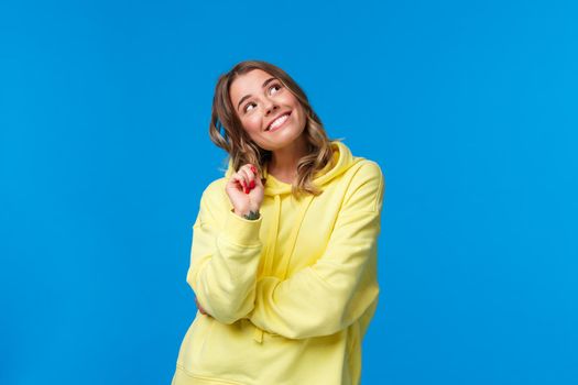 Dreamy silly young caucasian woman with blond hair, looking up thoughtful fantasizing, imaging something with curious smile, thinking or dreaming, standing blue background romantic.