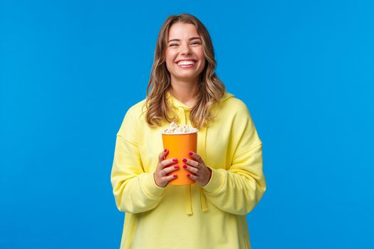 Leisure, fun and youth concept. Friendly happy blond european woman enjoying weekends, eating popcorn and watching comedy, visit cinema to see new movie, stand blue background.