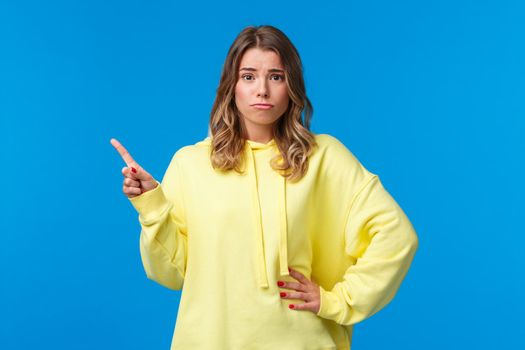 Upset and insecure cute european blond young woman in yellow hoodie, having doubts, being skeptical about topic, pointing finger upper left corner and look unsure camera, blue background.