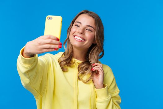 Close-up portrait tender cute blond girl in yellow hoodie, holding mobile phone, taking selfie with smartphone posing adding photo filter in appllication, standing blue background.