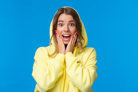 Close-up portrait of impressed and fascinated blond girl in hood react to amazing great news, touch cheeks gasping and smiling wondered, hear she won prize, standing blue background.