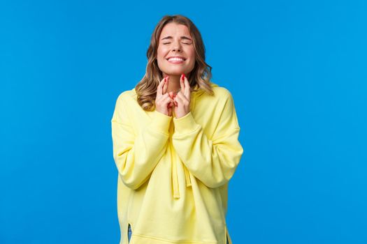 Wishing all well. Hopeful optimistic blond girl in yellow hoodie praying god make wish come true, cross fingers good luck pleading with closed eyes, supplicating, stand blue background.