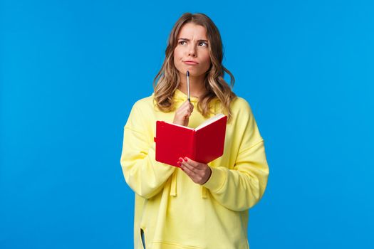 Waist-up portrait of cute european blond female in yellow hoodie making plans, solving homework or write diary, touching lip with pen pondering look upper left corner thoughtful, hold red notebook.