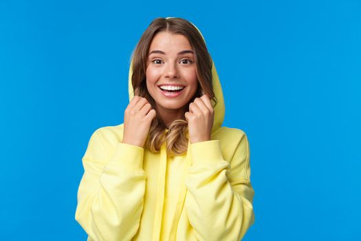 Lifestyle, people and youth concept. Close-up portrait of beautiful smiling blond female put on hoodie, looking camera with upbeat enthusiastic expression, standing blue background.