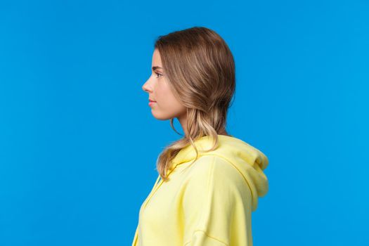 Close-up profile portrait of serious-looking attractive young caucasian female with short blond hair, looking left, standing in yellow hoodie, standing blue background straight.