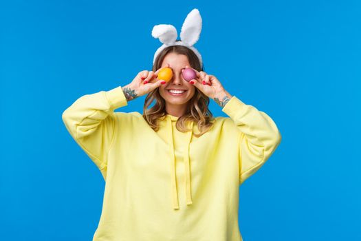 Holidays, traditions and celebration concept. Happy carefree smiling caucasian woman celebrating Easter, holding two painted eggs on eyes and grinning, wear cute rabbit ears, blue background.