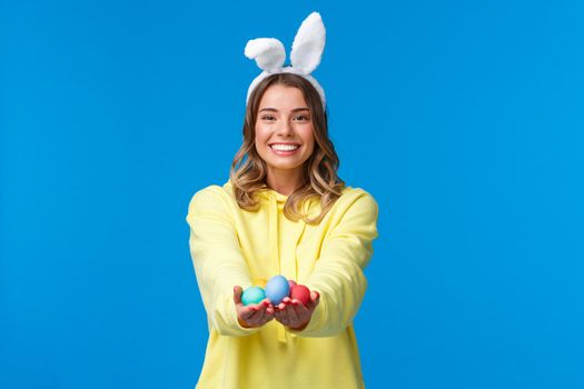 Holidays, people and fun concept. Charismatic cute blond girl holding coloured Easter eggs, wearing bunny ears and smiling joyfully, congratulate family with holy day, blue background.