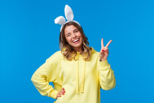 Holidays, traditions and celebration concept. Lovely and tender blond girl in yellow hoodie, tilt head kawaii wink and smiling as wearing rabbit ears, show peace gesture for goodwill, happy easter.