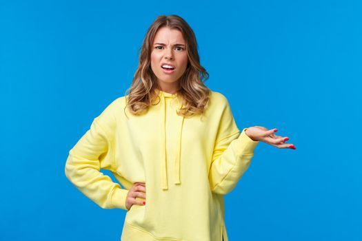 Whats wrong, why. Confused and frustrated blond girl dealing with person complains, shrugging and raise one hand in dismay grimacing perplexed, look camera questioned, blue background.