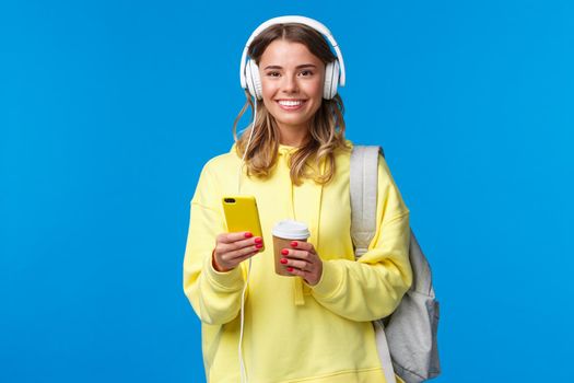 Education, lifestyle and teenagers concept. Cheerful young blond girl in headphones listening music and texting friends while waiting for them near cafe, drinking take-away coffee hold backpack.