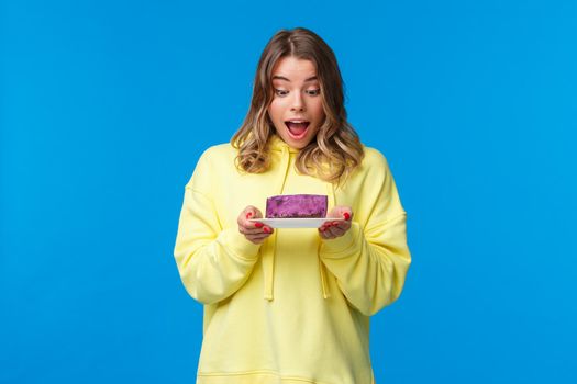 Celebration, party and lifestyle concept. Happy and pleased blond caucasian girl in yellow hoodie, looking fascinated at tasty berry cake with desire, want have bite, stand blue background.