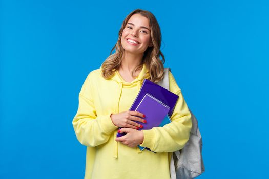 Charismatic joyful european female carry backpack and notebooks as going next lecture, smiling happy, enjoying student lifestyle, studying in university, standing blue background.
