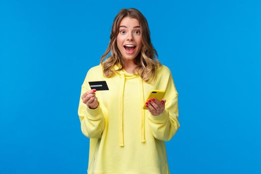 Amused and surprised cute blond girl receive cool cashback or banking offer after using new credit card with special student offer, holding mobile phone and smiling camera.