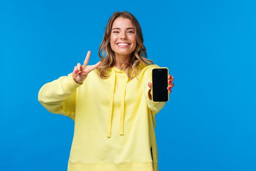 Cute smiling happy blond girl in yellow hoodie showing you her online blog or social media page on mobile phone display, make peace gesture and grinning, stand blue background.